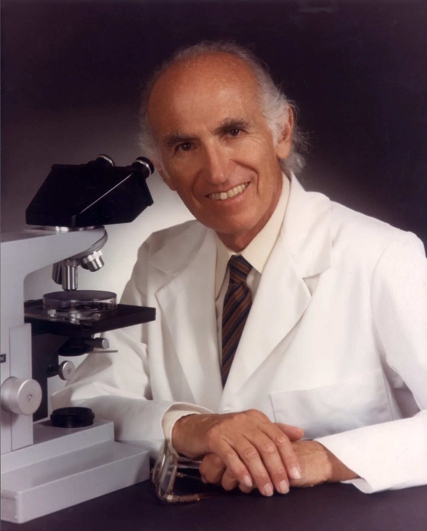 Dr. Jonas Salk in a white lab coat, seated beside a microscope